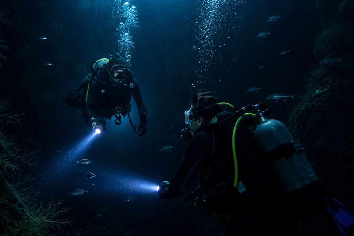 Night diving in the Galápagos with two scuba divers illuminating fish and reef life during our agency’s cruise excursion